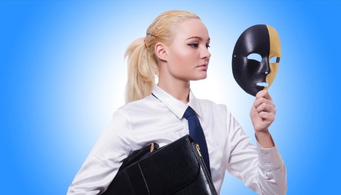 A blond woman wearing a blue tie and holding a mask in front of her face